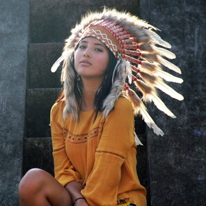May include: A woman wearing a brown and white feathered headdress with red and white beads. She is wearing a yellow shirt with a lace design.