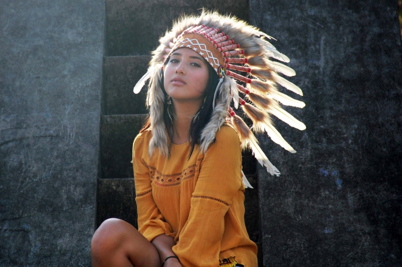 May include: A woman wearing a brown and white feathered headdress with red and white beads. She is wearing a yellow shirt with a lace design.