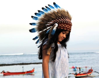 Puede incluir: Una persona joven con un tocado de plumas azules y marrones con una banda de patrón marrón y blanco. El tocado tiene una franja de plumas alrededor de la parte inferior. La persona lleva una camisa azul claro y tiene el pelo largo y oscuro.