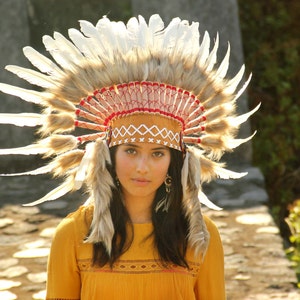 May include: A woman wearing a brown and white feathered headdress with red and white trim. She is wearing a yellow shirt with a brown and white pattern.