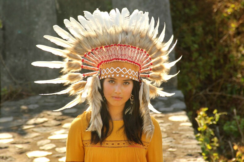 May include: A woman wearing a brown and white feathered headdress with red and white trim. She is wearing a yellow shirt with a brown and white pattern.