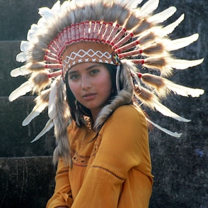 May include: A woman wearing a brown and white feathered headdress with red and white accents. She is wearing a yellow blouse and sitting on a set of stairs.