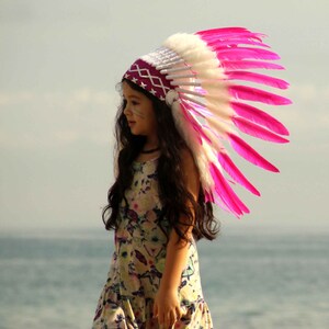 May include: A young girl wearing a pink and white feathered headdress and a floral dress. She is standing on a sandy beach with the ocean in the background.