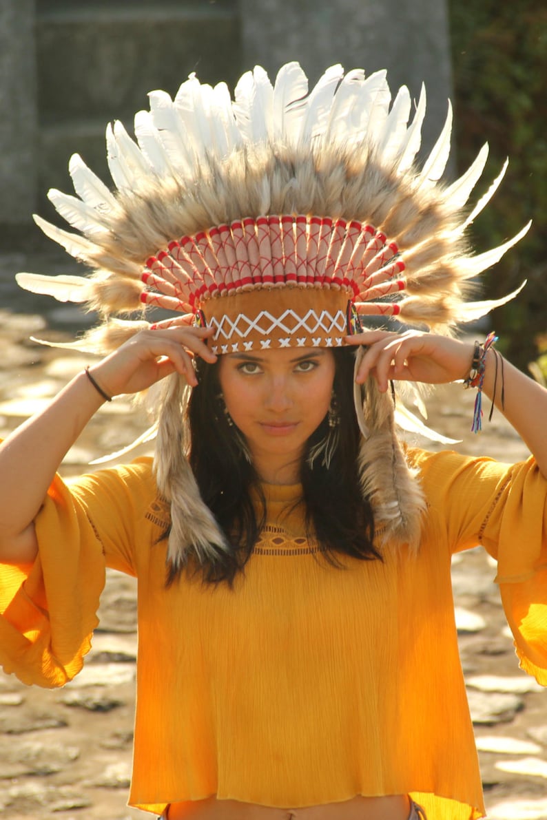May include: A woman wearing a brown and white feathered headdress with red and white trim. She is wearing a yellow top and has her hands on the headdress.