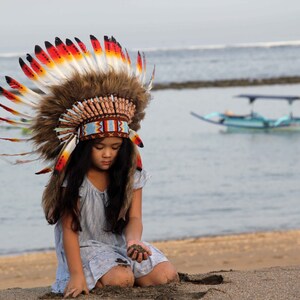 Puede incluir: Una persona joven con un tocado nativo americano colorido con plumas y cuentas. Est&aacute; arrodillada en una playa de arena con el oc&eacute;ano de fondo.