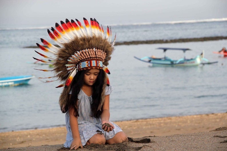 Puede incluir: Una persona joven con un tocado nativo americano colorido con plumas y cuentas. Est&aacute; arrodillada en una playa de arena con el oc&eacute;ano de fondo.