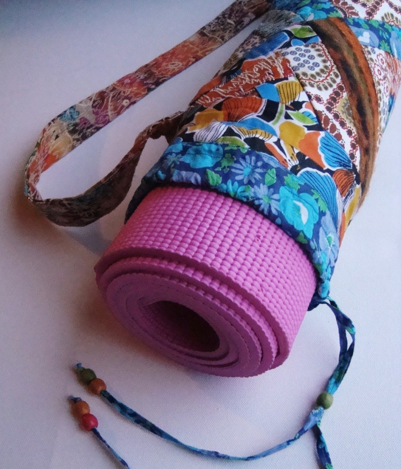 May include: A pink yoga mat rolled up and secured in a colorful fabric carrying case with a long strap. The case has a floral pattern with blue, orange, and brown accents.