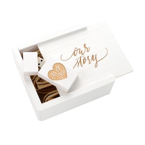 Puede incluir: Caja de madera blanca con una tapa que dice "our story" en letras doradas. La caja está abierta y contiene una unidad USB de madera con forma de corazón con las palabras "Mr & Mrs" grabadas.