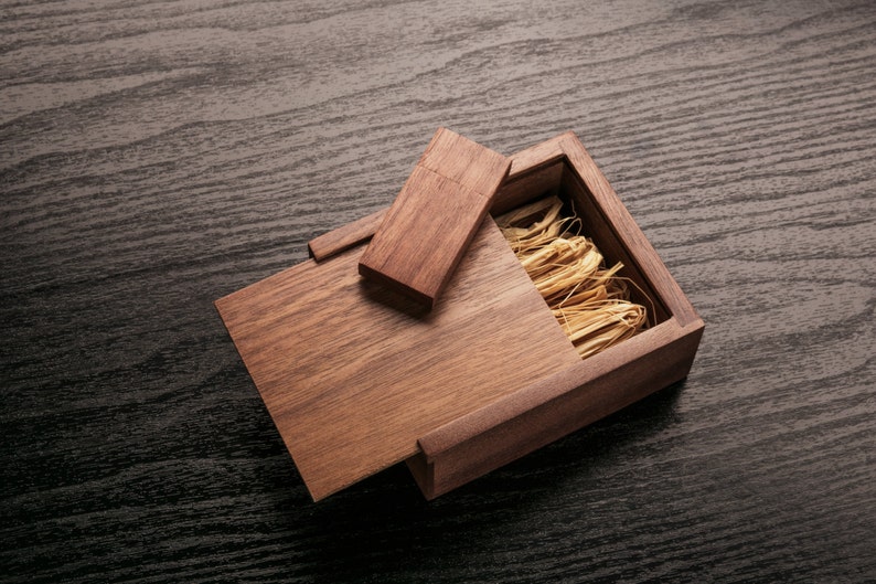 May include: A wooden USB flash drive and its matching box, both crafted from a warm-toned wood. The box is open, revealing a bed of straw-like material. The lid of the box is partially open, and the flash drive rests on top.