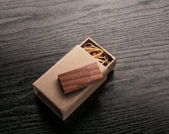 Puede incluir: Una memoria USB de madera en una pequeña caja de cartón con relleno de paja. La caja está abierta y la memoria USB es visible.