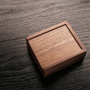 May include: A rectangular wooden box with a lid, crafted from light brown wood. The box is set at an angle on a dark, textured surface, showcasing the natural wood grain and the box's construction.