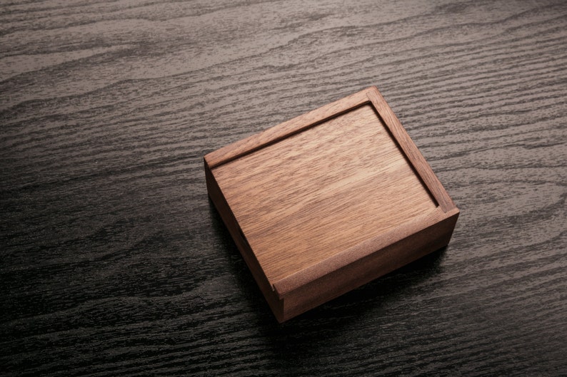 May include: A rectangular wooden box with a lid, crafted from light brown wood. The box is set at an angle on a dark, textured surface, showcasing the natural wood grain and the box's construction.