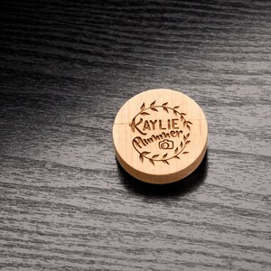 May include: A wooden round disc with the text "Kaylie Summer" and a camera icon engraved on it. The disc is surrounded by a wreath of leaves.