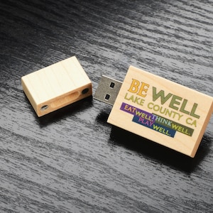 May include: Wooden USB drive with a light brown finish. The drive has a rectangular shape and features the text "BE WELL LAKE COUNTY CA EAT WELL THINK WELL PLAY WELL."