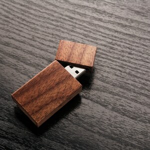 May include: A wooden USB flash drive with a natural wood grain finish. The USB drive is partially open, revealing the silver connector. The wooden case is rectangular and has a hinged lid. The background is a dark, textured surface.