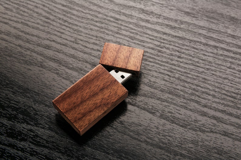 May include: A wooden USB flash drive with a natural wood grain finish. The USB drive is partially open, revealing the silver connector. The wooden case is rectangular and has a hinged lid. The background is a dark, textured surface.
