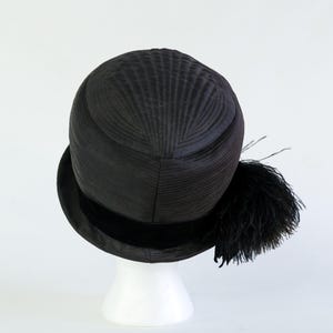 Puede incluir: Un sombrero cloche negro con una corona texturizada y un ala ancha. El sombrero presenta una banda de terciopelo negro y un adorno decorativo de plumas negras. El sombrero se exhibe sobre un soporte blanco.