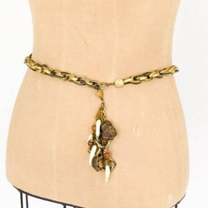 May include: A gold-toned chain belt with a decorative pendant. The belt features a series of interlocking links and a dangling charm with brown, white, and gold accents. The pendant includes beads and leaf-like shapes.