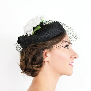 May include: A black pillbox hat with a white flower and a black veil. The hat is worn on a person's head.