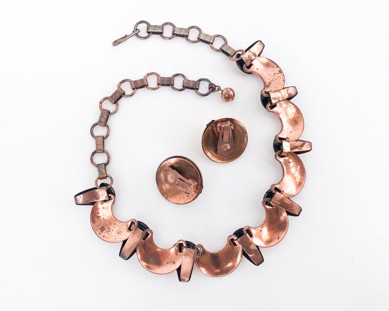 May include: A copper necklace with a chain and a matching pair of earrings. The necklace is made of a series of crescent-shaped copper pieces with black accents.