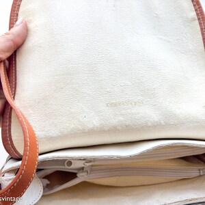 Puede incluir: Bolso de mano de cuero color crema con ribete marr&oacute;n y correa beige. El interior tiene un bolsillo con cremallera y forro crema. El bolso est&aacute; abierto, mostrando el interior.