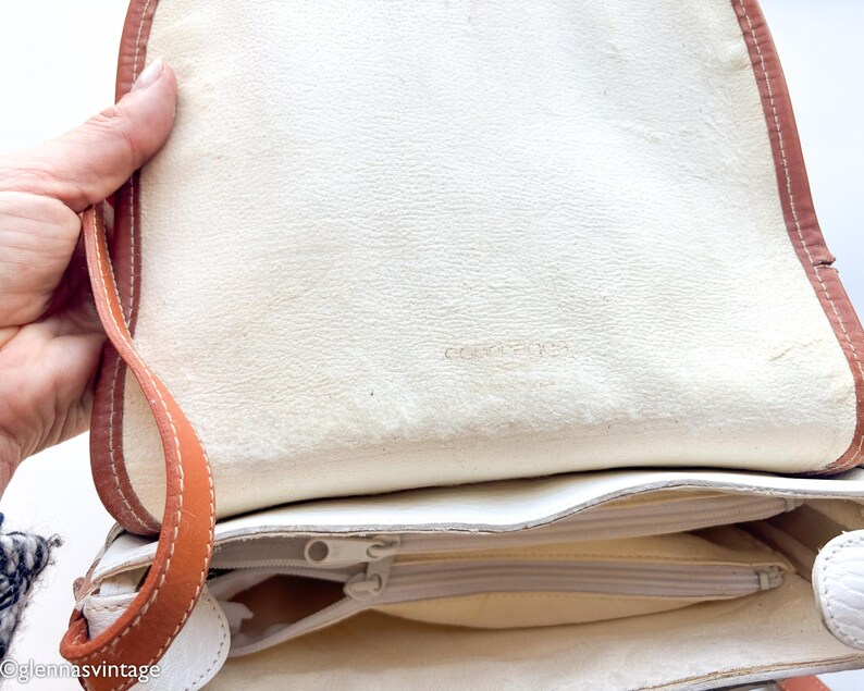 Puede incluir: Bolso de mano de cuero color crema con ribete marr&oacute;n y correa beige. El interior tiene un bolsillo con cremallera y forro crema. El bolso est&aacute; abierto, mostrando el interior.