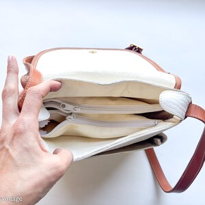Puede incluir: Vista abierta de un bolso de hombro vintage con interior blanco y m&uacute;ltiples compartimentos con cremallera. El bolso presenta una correa y ribete de cuero marr&oacute;n, con una solapa blanca y un cierre dorado. El bolso est&aacute; abierto, revelando el interior.