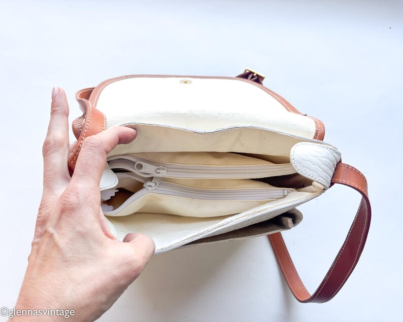Puede incluir: Vista abierta de un bolso de hombro vintage con interior blanco y m&uacute;ltiples compartimentos con cremallera. El bolso presenta una correa y ribete de cuero marr&oacute;n, con una solapa blanca y un cierre dorado. El bolso est&aacute; abierto, revelando el interior.