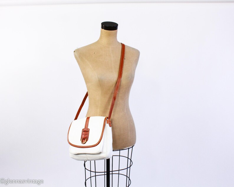 Puede incluir: Un bolso bandolera blanco con una correa y detalles en cuero marr&oacute;n. El bolso tiene un cierre de solapa con una correa de cuero marr&oacute;n y una hebilla dorada. El bolso se exhibe en un maniqu&iacute;.