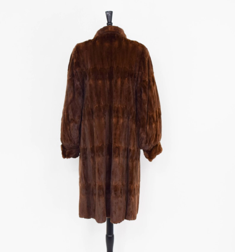 May include: A long brown fur coat with a collar and long sleeves. The coat is on a mannequin and is displayed against a white background.