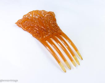 May include: An amber-coloured hair comb with an ornate, openwork design at the top. The comb features five tines and a gradient from amber to a lighter, almost translucent colour at the tips. The comb is likely vintage.