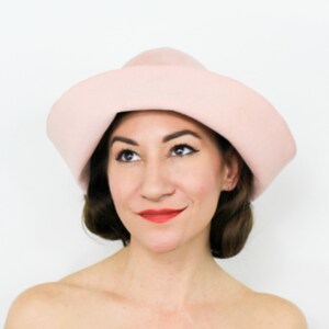 May include: A woman wearing a light pink felt hat with a wide brim. The hat has a unique shape, with a pointed crown and a curved brim.