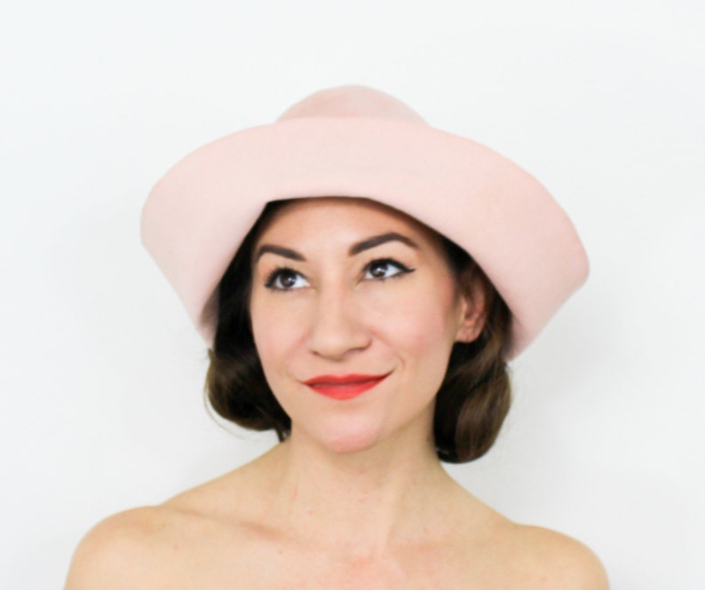 May include: A woman wearing a light pink felt hat with a wide brim. The hat has a unique shape, with a pointed crown and a curved brim.