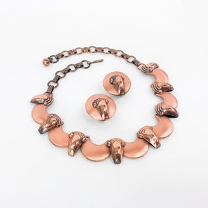 May include: A copper necklace and earrings set with a scalloped design. The necklace has a chain and a row of crescent-shaped pendants with a raised design. The earrings are simple studs with a similar design.
