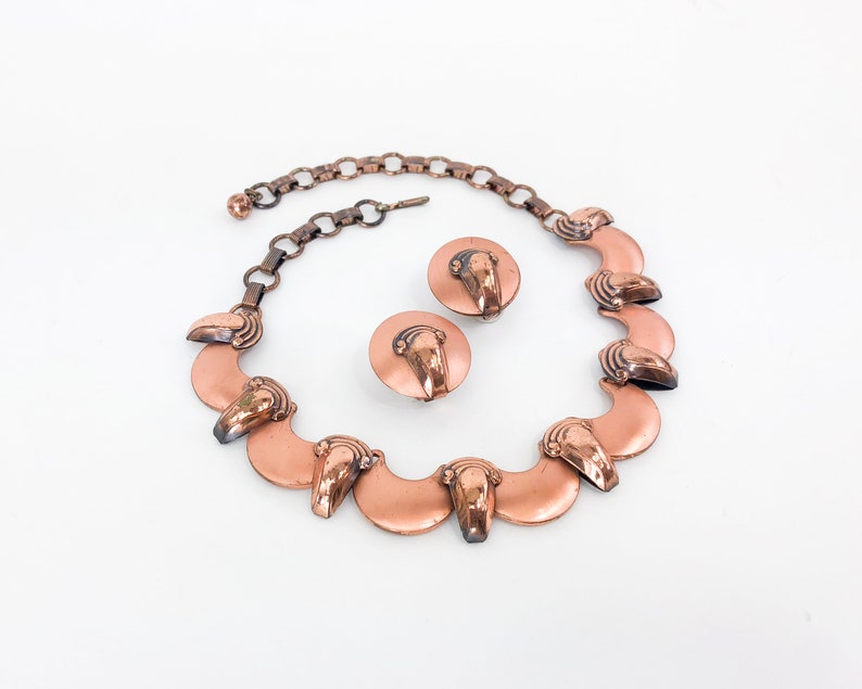 May include: A copper necklace and earrings set with a scalloped design. The necklace has a chain and a row of crescent-shaped pendants with a raised design. The earrings are simple studs with a similar design.