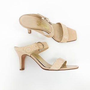 May include: A pair of beige heeled sandals with a buckle strap and the Salvatore Ferragamo logo embossed on the leather.
