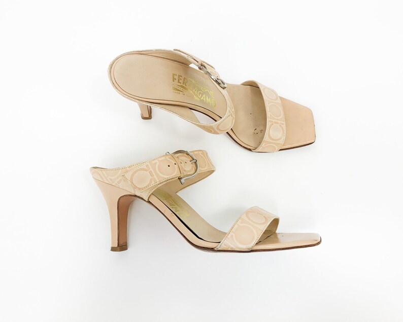 May include: A pair of beige heeled sandals with a buckle strap and the Salvatore Ferragamo logo embossed on the leather.