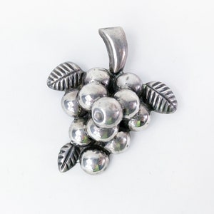 May include: A silver-toned grape cluster brooch with detailed leaves and a stem. The brooch features a bunch of round, polished grapes. The leaves have detailed veining. The brooch is set against a white background.