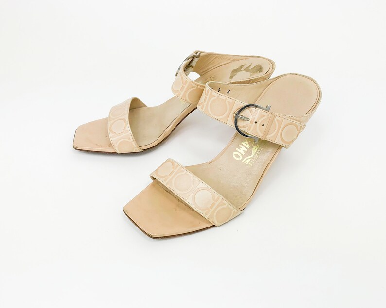 May include: A pair of beige leather heeled sandals with a buckle closure and a Salvatore Ferragamo logo on the sole.