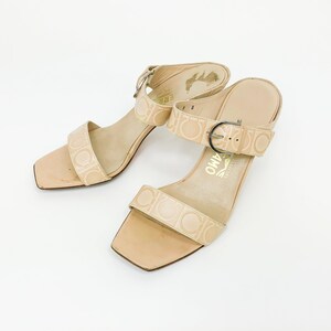 May include: A pair of beige heeled sandals with a buckle strap and a logo pattern. The sandals have a square toe and a low heel.