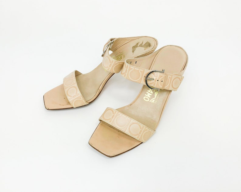 May include: A pair of beige heeled sandals with a buckle strap and a logo pattern. The sandals have a square toe and a low heel.