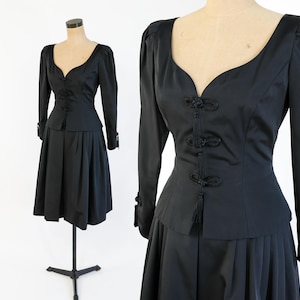 Peut inclure: Ensemble de robe deux pièces noire vintage. Le haut à manches longues présente un décolleté en V et des boutons décoratifs. La jupe est une conception plissée jusqu'aux genoux. La robe est présentée sur un mannequin.