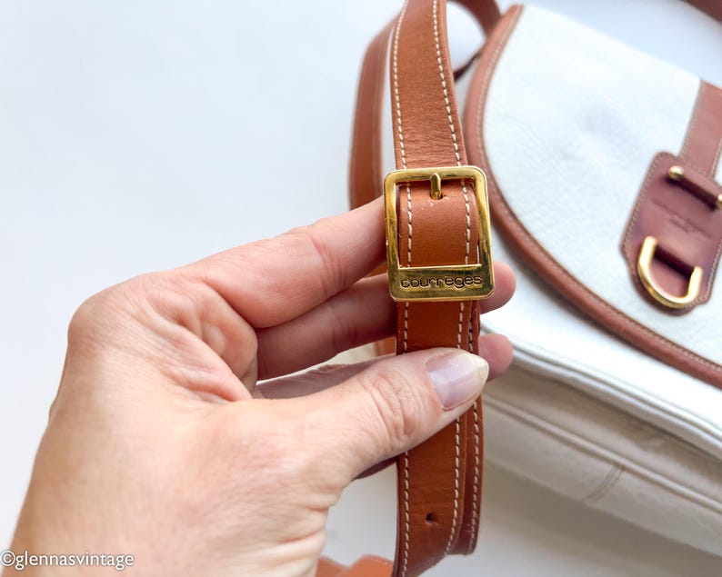 Puede incluir: Bolso Courreges vintage con cuerpo blanco y detalles en cuero marr&oacute;n. El bolso presenta una hebilla y una correa doradas, con el logotipo de Courreges visible. El bolso est&aacute; sostenido por una mano.
