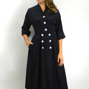 Pode incluir: Um vestido midi azul marinho com gola e uma dupla fileira de bot&otilde;es brancos na frente. O vestido tem um corpete justo e uma saia rodada.