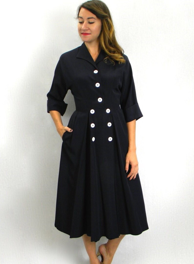 Pode incluir: Um vestido midi azul marinho com gola e uma dupla fileira de bot&otilde;es brancos na frente. O vestido tem um corpete justo e uma saia rodada.