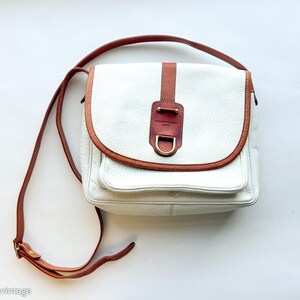 Puede incluir: Bolso bandolera de cuero blanco con ribetes y correa de cuero marr&oacute;n. El bolso tiene un cierre de solapa con un adorno de cuero marr&oacute;n y una hebilla de metal dorado. Forma rectangular con un bolsillo frontal.