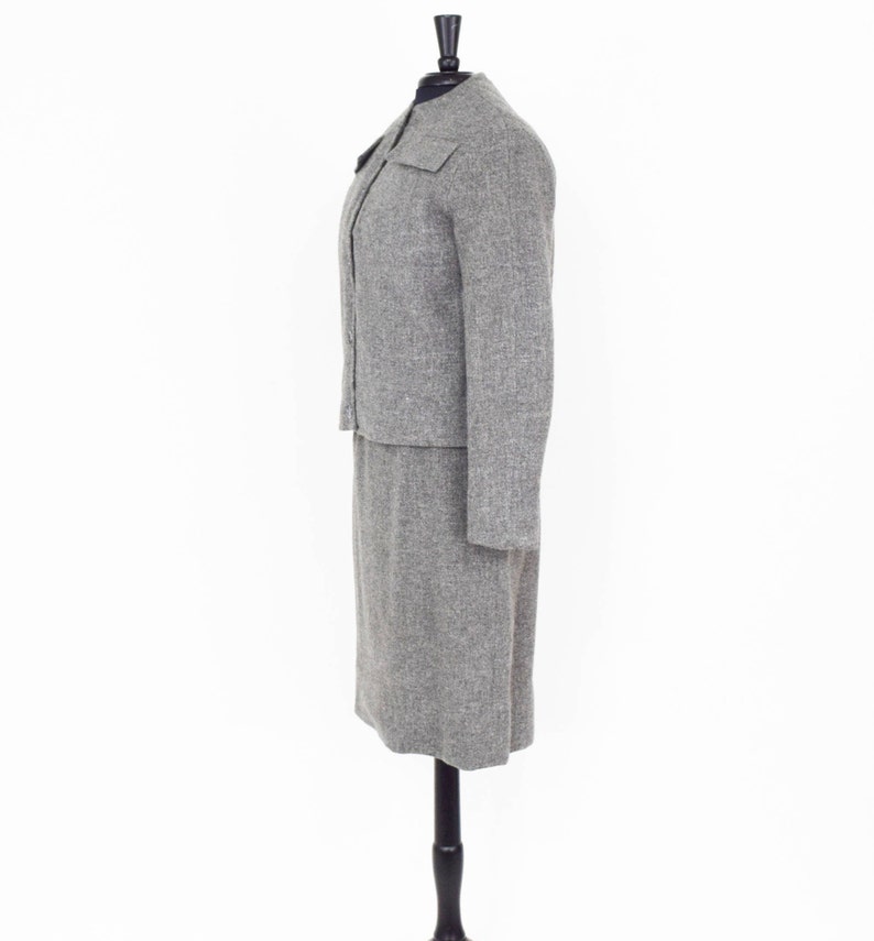 May include: A gray tweed skirt suit with a long skirt and a fitted jacket. The jacket has a mandarin collar and a single button closure.