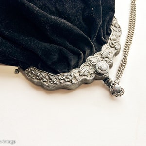 May include: A vintage black velvet purse with a silver chain and ornate metal clasp. The clasp features floral designs and a small round button.