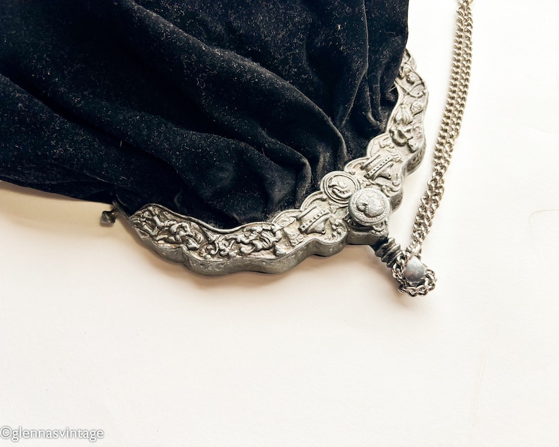 May include: A vintage black velvet purse with a silver chain and ornate metal clasp. The clasp features floral designs and a small round button.
