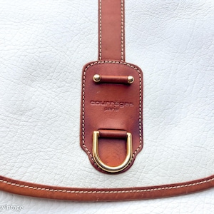 Puede incluir: Primer plano de un bolso de mano de cuero blanco con detalles en cuero marr&oacute;n. El bolso presenta una correa de cuero marr&oacute;n y una hebilla de metal dorada. El nombre de la marca "courr&egrave;ges paris" es visible en la correa.